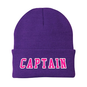 May include: A purple beanie with the word "CAPTAIN" in bright pink block letters outlined in white. The beanie is a solid color and has a folded brim.