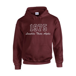 May include: A maroon hooded sweatshirt with a front pocket and a drawstring hood. The sweatshirt features the year "1975" in white block letters, with "Lambda Theta Alpha" in cursive script underneath. The Gildan logo is visible on the tag.