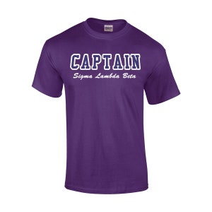 May include: A purple t-shirt with the word "CAPTAIN" in white block letters with a blue outline. Below the word "CAPTAIN" is the text "Sigma Lambda Beta" in white script. The shirt is made of a soft material.