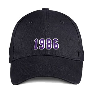 Puede incluir: Una gorra de b&eacute;isbol negra con el n&uacute;mero "1986" bordado en morado en la parte delantera.