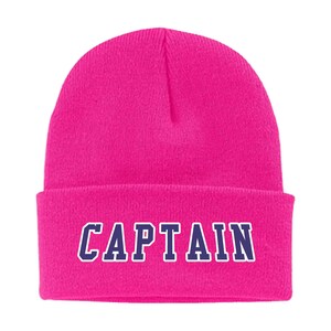 May include: A vibrant pink beanie with the word "CAPTAIN" in white outlined, navy blue letters. This warm winter hat is a stylish accessory for cold weather.