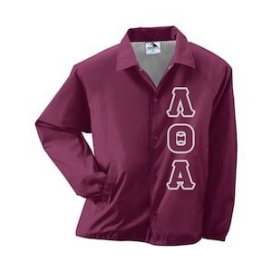 May include: A maroon coach jacket with white lettering spelling out "ΛOA" vertically down the front. The jacket has a button-up closure, a collar, and two front pockets. The jacket is made of a lightweight material.
