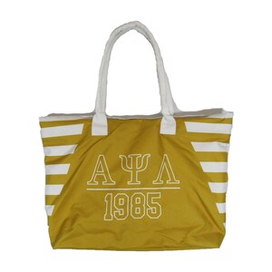 May include: Mustard yellow tote bag with white horizontal stripes and white handles. The bag features the Greek letters A, Ψ, Λ, and the number 1985 in white block letters. The tote bag is designed for carrying items.