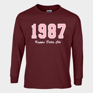 May include: A maroon long-sleeve shirt with the year "1987" in pink block letters outlined in white. Below the numbers, the text "Kappa Delta Chi" is written in a cursive font. The shirt has a crew neck and ribbed cuffs.