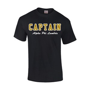 May include: Black crew neck t-shirt with the word "CAPTAIN" in gold block letters with a white outline. Below the word "CAPTAIN" is the text "Alpha Phi Lambda" in a cursive font.