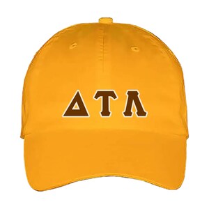 May include: Mustard yellow baseball cap with the Greek letters Delta, Tau, and Lambda in brown. The letters are outlined in white. The cap has a curved brim and an adjustable strap at the back.