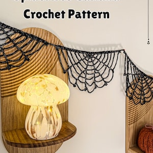 May include: A black crochet spiderweb garland featuring a repeating web design, displayed above a mushroom-shaped lamp. The text "Spiderweb Garland Crochet Pattern" is printed at the top of the image in a bold, black, sans-serif font.