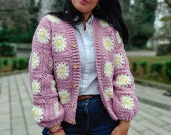 May include: A pink crocheted cardigan with white daisy flowers. The cardigan has a button-up front and long sleeves.