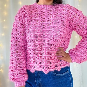 Könnte beinhalten: Ein rosa gestrickter Pullover mit Blumenmuster. Der Pullover hat einen runden Ausschnitt und lange Ärmel.