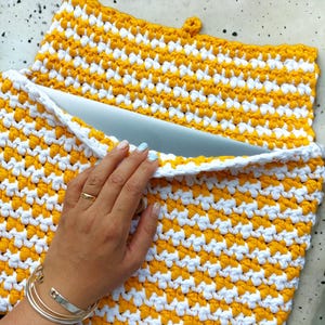 Può includere: Una custodia per laptop in crochet giallo e bianco con chiusura a zip. La custodia è realizzata con una maglia testurizzata e ha un design semplice.