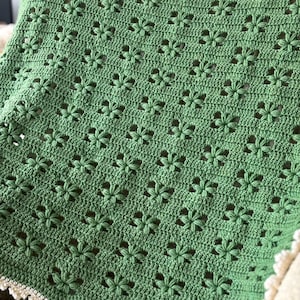 Puede incluir: Una manta verde hecha a mano con un patrón floral. La manta está hecha de hilo suave y tiene un diseño calado delicado. Los bordes están acabados con un borde festoneado color crema. La manta está doblada sobre una superficie de color claro.