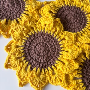 May include: Handmade crochet sunflower coasters in yellow and brown. The coasters feature a detailed stitch pattern, mimicking the petals and centre of a sunflower. The bright yellow petals contrast with the brown centre, creating a cheerful, decorative item.