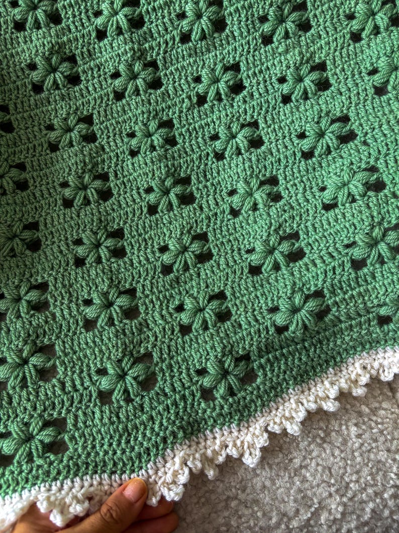 a crochet baby blanket made out of sage wool yarn lying on a sofa. the blanket features a texture flower puff stitch all along and a crowned picot border in white