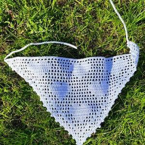 May include: A white crocheted triangular scarf with a delicate openwork pattern. The scarf is laid out on a bed of green grass.