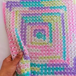 Può includere: Una copertina per neonati all'uncinetto con un motivo quadrato in tonalità di rosa, viola, verde e bianco.