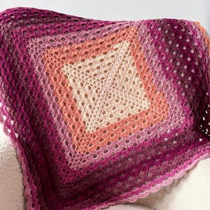May include: A handmade crochet blanket in a square shape, featuring a gradient of colors from a deep magenta to a soft cream center. The blanket has an open, lacy design with a scalloped edge, perfect for home decor.