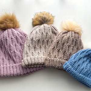 May include: Four knitted beanies in shades of purple, tan, and blue, each topped with a faux fur pom-pom. The hats have a textured, ribbed design. The beanies are a cozy winter accessory.