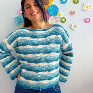 Könnte beinhalten: Ein langärmeliger Häkelpullover mit einem wellenförmigen, horizontalen Streifenmuster in den Farben Petrol, Hellblau und Creme. Das Kleidungsstück hat einen weiten, runden Ausschnitt und eine lockere Passform mit einem strukturierten Maschenbild.