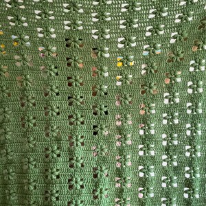 Op de afbeelding: Een handgemaakte, groene gehaakte deken met een bloemenpatroon. De deken heeft een herhalend ontwerp van kleine, driedimensionale bloemen, waardoor een textuur ontstaat. De bovenrand heeft een cr&egrave;mekleurige, geschulpte rand.