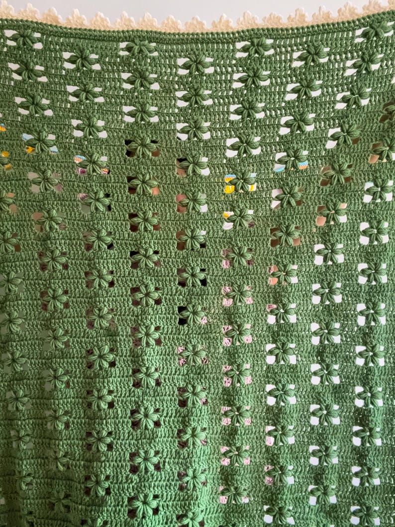 Op de afbeelding: Een handgemaakte, groene gehaakte deken met een bloemenpatroon. De deken heeft een herhalend ontwerp van kleine, driedimensionale bloemen, waardoor een textuur ontstaat. De bovenrand heeft een cr&egrave;mekleurige, geschulpte rand.