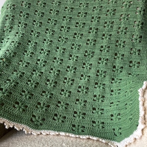 Op de afbeelding: Een groene gehaakte deken met een bloemmotief, over een stoel gedrapeerd. De deken heeft een witte, geschulpte rand. Gemaakt van zacht garen, geschikt voor een baby of als decoratief kleed. Afmetingen: ongeveer 120 cm x 150 cm.