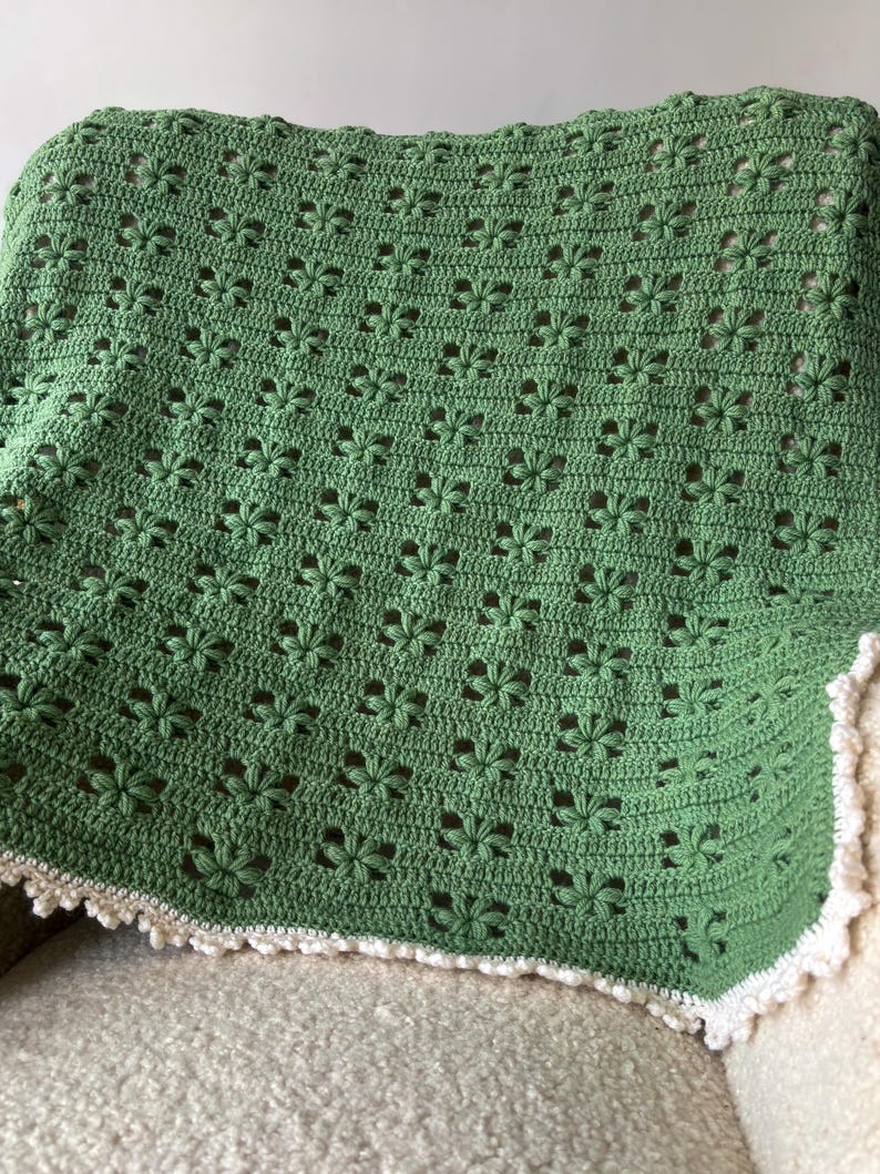 Op de afbeelding: Een groene gehaakte deken met een bloemmotief, over een stoel gedrapeerd. De deken heeft een witte, geschulpte rand. Gemaakt van zacht garen, geschikt voor een baby of als decoratief kleed. Afmetingen: ongeveer 120 cm x 150 cm.