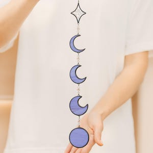 May include: A hanging stained glass moon phase decoration. The phases include a star, three crescent moons, and a full moon, all in shades of purple. The phases are connected by a silver chain and small beads.