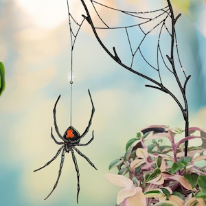 May include: Black spider decoration with an orange marking on its back, suspended from a black web structure. The spider hangs in front of a window, with plants in the foreground. The web is attached to a branch-like structure.