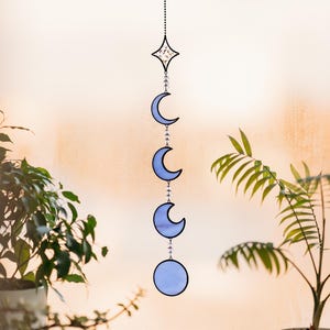 Op de afbeelding: Een zonneweergave met een hemels thema, met een ster en maanfasen in blauwe tinten. Het decoratieve stuk is opgehangen aan een zuignap, met een ketting van kralen die de elementen verbindt. De zonneweergave wordt tentoongesteld voor een raam, met planten op de voorgrond.