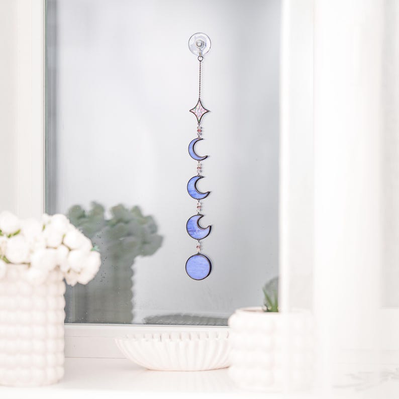 Op de afbeelding: Een raamhanger met een hemels thema, met een ster en maanfasen in blauw en roze. Het decoratieve stuk wordt opgehangen met een ketting en een zuignap. Een uniek woondecoratie-item dat een vleugje fantasie toevoegt. Afmetingen in centimeters.