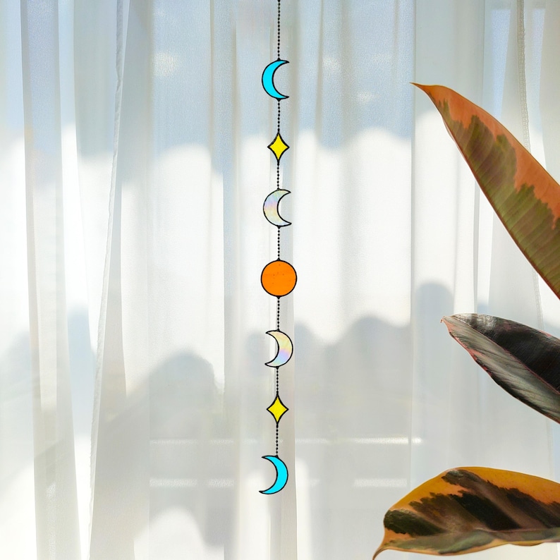 Op de afbeelding: Een hangende mobiel met hemelse vormen: halve manen, een ruit en een cirkel. De vormen zijn in blauw, geel en oranje, verbonden door een ketting. De achtergrond is een witte, transparante gordijn, wat een zacht licht cre&euml;ert.