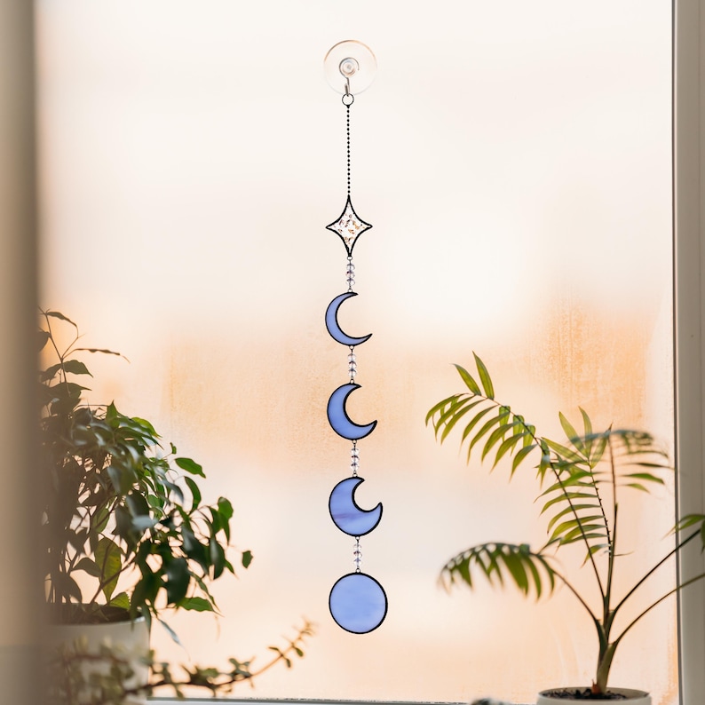 Op de afbeelding: Een zonneweergave met een hemels thema, met een ster en maanfasen in blauwe tinten. Het decoratieve stuk is opgehangen aan een zuignap, met een ketting van kralen die de elementen verbindt. De zonneweergave wordt tentoongesteld voor een raam, met planten op de voorgrond.