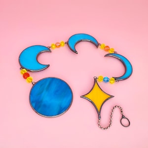 Op de afbeelding: Een gebrandschilderde zonnevanger met een blauwe maan, een gele ster en een blauwe cirkel, allemaal verbonden met kralen. De zonnevanger heeft een metalen rand en een ketting. De achtergrond is roze.