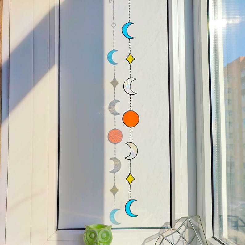 Op de afbeelding: Een decoratieve zonnevanger met maanfasen en sterren. Het heeft blauwe, gele, oranje en zilverkleurige glazen vormen. De zonnevanger hangt voor een raam. Een groen uilornament is zichtbaar aan de onderkant.