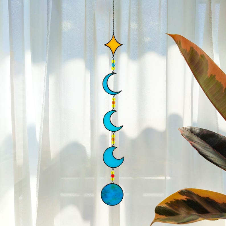 Op de afbeelding: Een hemels thema zonnevanger met een gele ster, drie blauwe halve manen en een blauwe bol, verbonden door een kralenketting. Het decoratieve stuk hangt voor een transparant wit gordijn, met een plant op de achtergrond.