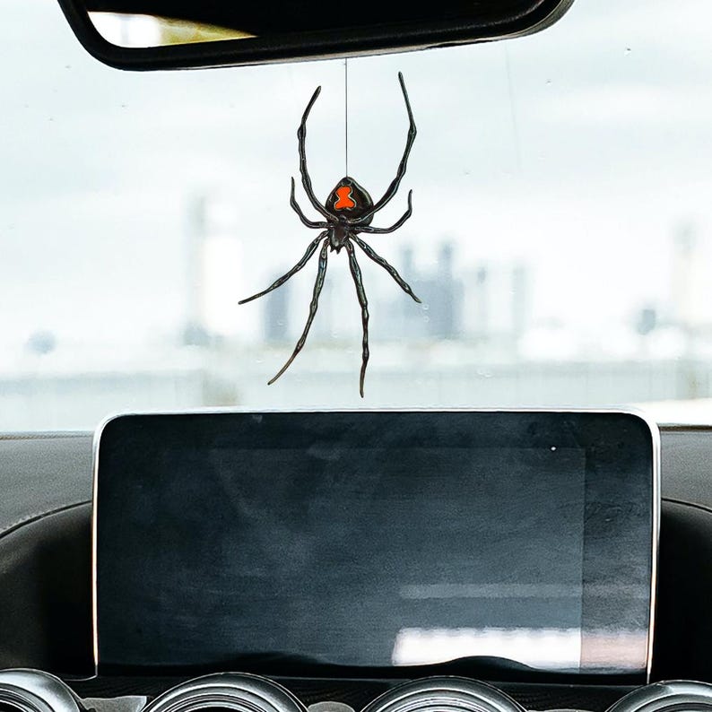 Op de afbeelding: Een zwarte weduwe spin auto charme hangend aan een touwtje. De spin heeft een rode zandloper markering op de rug. De charme hangt voor het dashboard en de achteruitkijkspiegel van een auto.