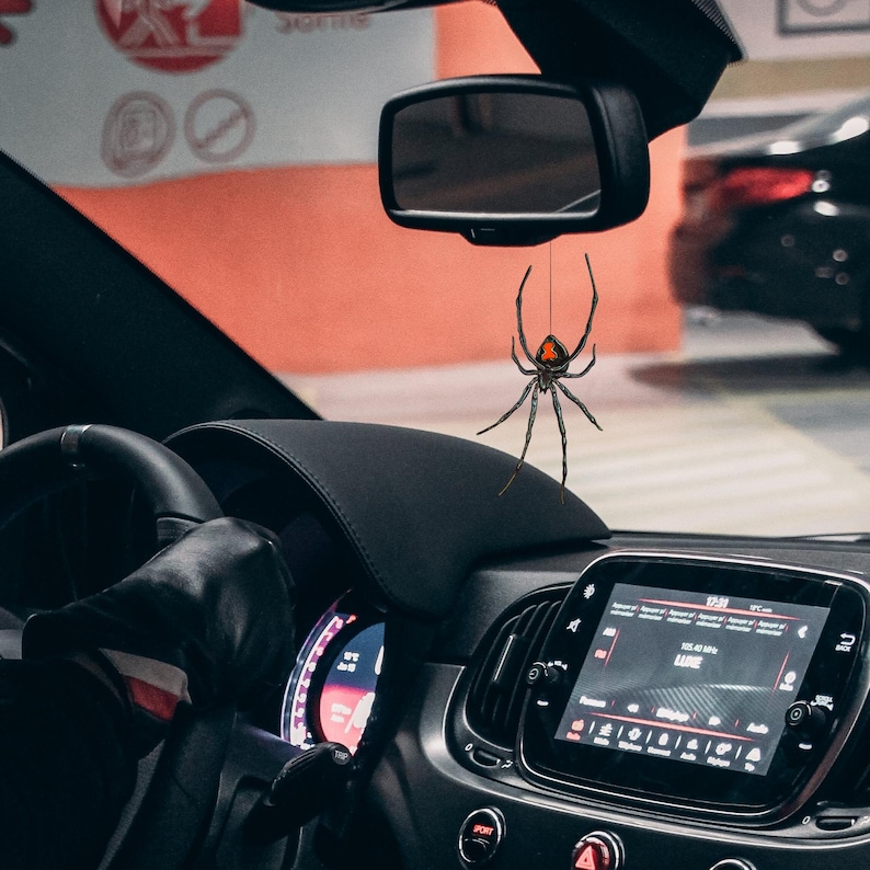 Op de afbeelding: Een zwarte spinnenauto-hanger met een rode markering op de rug hangt aan de achteruitkijkspiegel in een auto. Het dashboard en het stuur van de auto zijn zichtbaar, evenals een digitaal display met informatie.