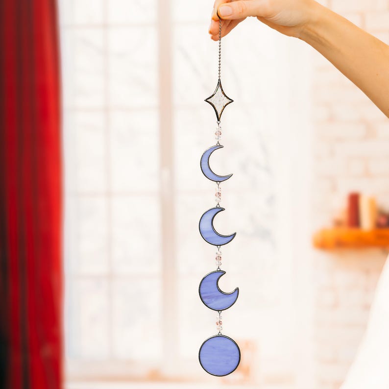 Op de afbeelding: Een hemels thema zonnevanger met een ster en maanfasen in blauw en helder glas. Het decoratieve stuk is opgehangen aan een ketting en versierd met kleine, roze kralen. Geschikt voor interieurdecoratie.