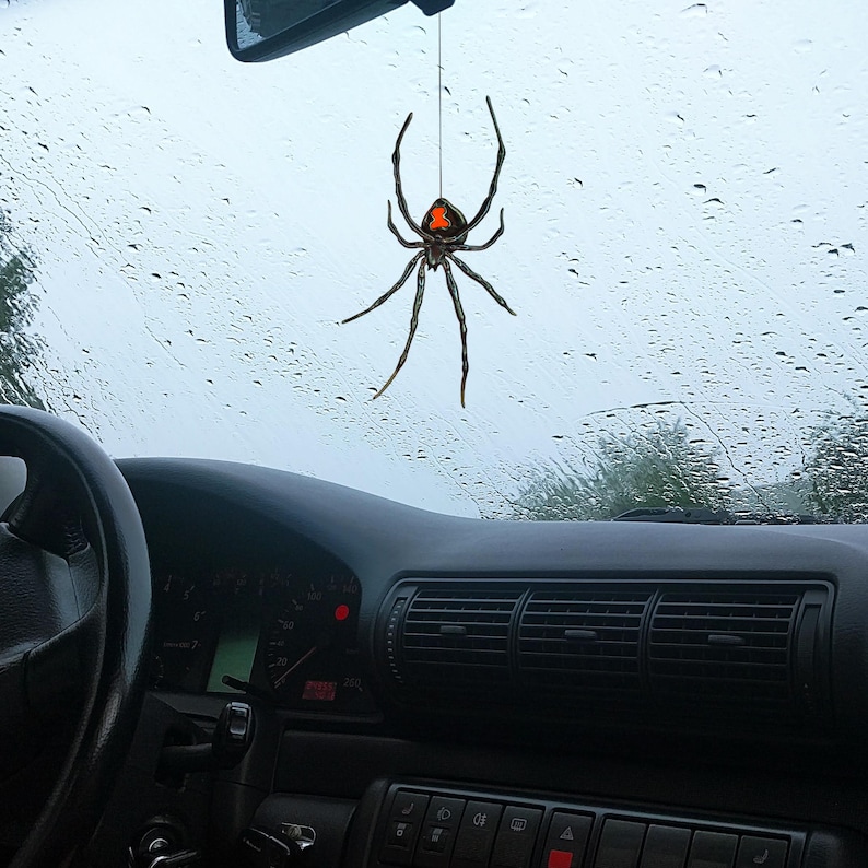 Op de afbeelding: Een zwarte spinnenauto-hanger met een rode zandlopermarkering hangt aan een touwtje voor een autoruit. De spin heeft lange poten en hangt tegen een achtergrond van een regenachtige dag.