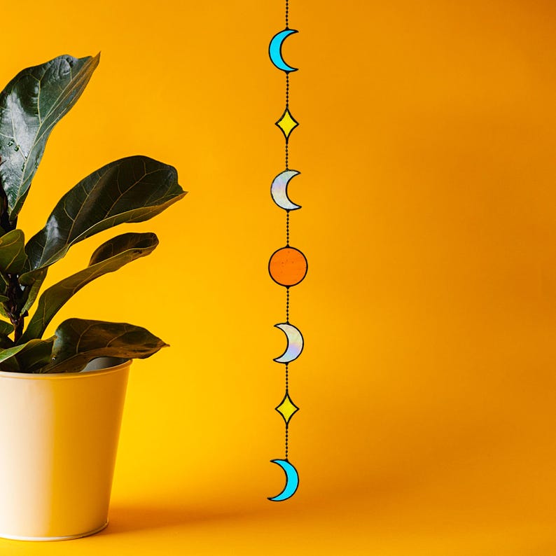 Op de afbeelding: Een wanddecoratie met een hemels thema, met een reeks gekleurde vormen: halve manen, een ruit en een cirkel, die aan elkaar zijn geregen. De kleuren zijn blauw, geel en oranje op een heldergele achtergrond. Een potplant is op de achtergrond te zien.