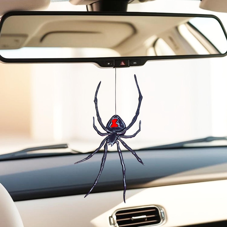 Op de afbeelding: Zwarte weduwe spin auto-hanger die aan een touwtje hangt. De spin heeft een zwart lichaam met een rode zandlopermarkering op de buik. De hanger hangt voor de achteruitkijkspiegel van een auto.