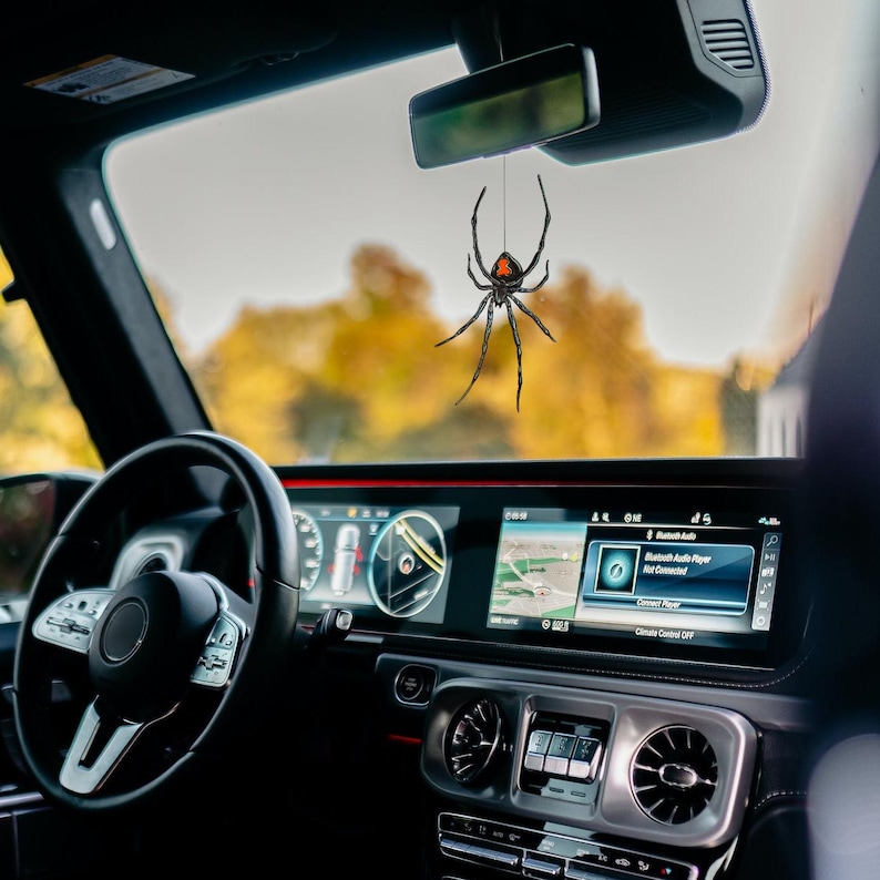 Op de afbeelding: Een zwarte en rode spinnen auto-hanger die aan de achteruitkijkspiegel van een auto hangt. De spin heeft een rode zandloper markering. Het interieur van de auto is zwart met een digitaal dashboard en stuur.