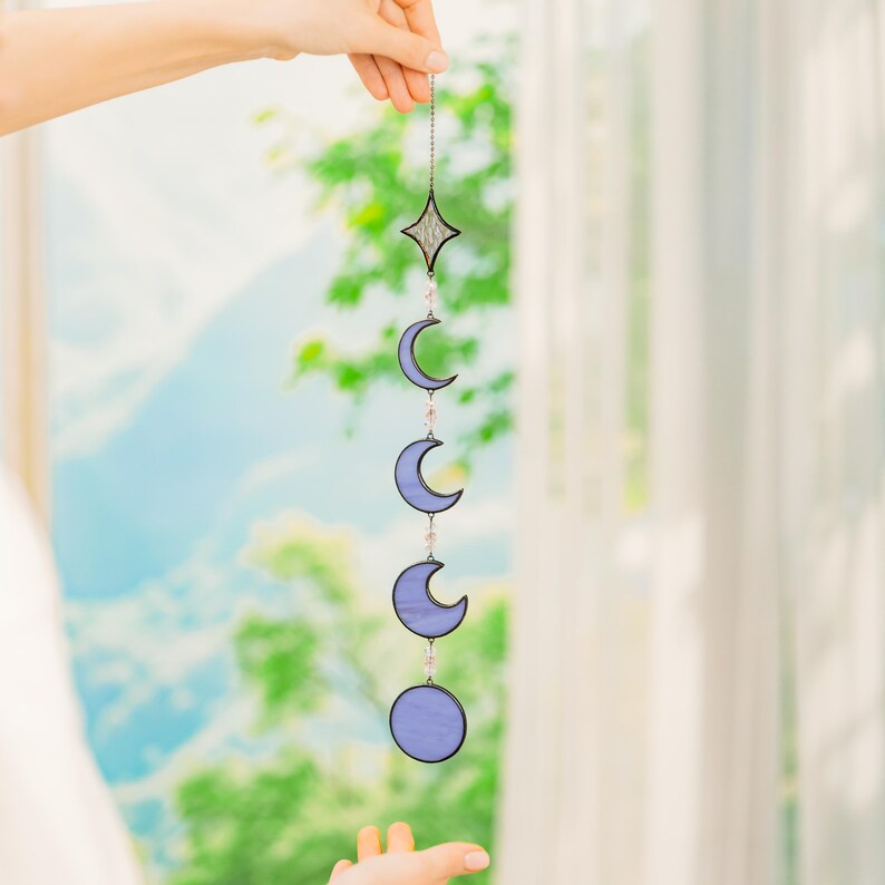 Op de afbeelding: Een hangende decoratie met maanfasen in paars, opgehangen aan een ketting. Een ster bekroont het ontwerp. Het object wordt door een hand vastgehouden.