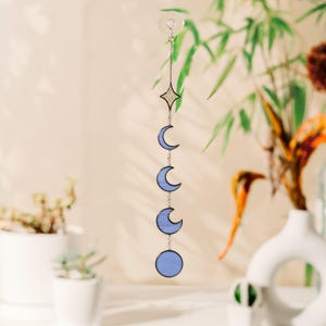 Op de afbeelding: Een hangende decoratie met een hemels thema, met maanfasen en een ster. De blauwe vormen zijn verbonden door een zilveren ketting en hangen aan een zuignap. Ontworpen om aan een raam of muur te hangen.