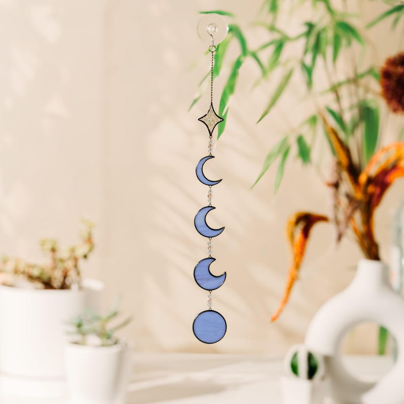 Op de afbeelding: Een hangende decoratie met een hemels thema, met maanfasen en een ster. De blauwe vormen zijn verbonden door een zilveren ketting en hangen aan een zuignap. Ontworpen om aan een raam of muur te hangen.