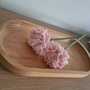 Könnte beinhalten: Zwei rosa gehäkelte Blumen mit grünen Stielen auf einem Holztablett.
