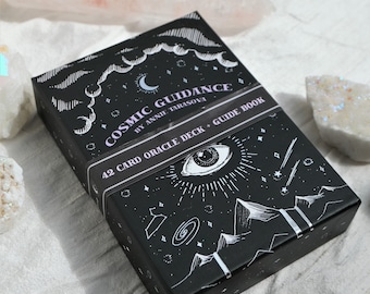May include: A black box with a white design of a celestial eye, stars, mountains, and clouds. The box is labeled "Cosmic Guidance by Annie Tarasova 42 Card Oracle Deck + Guide Book".