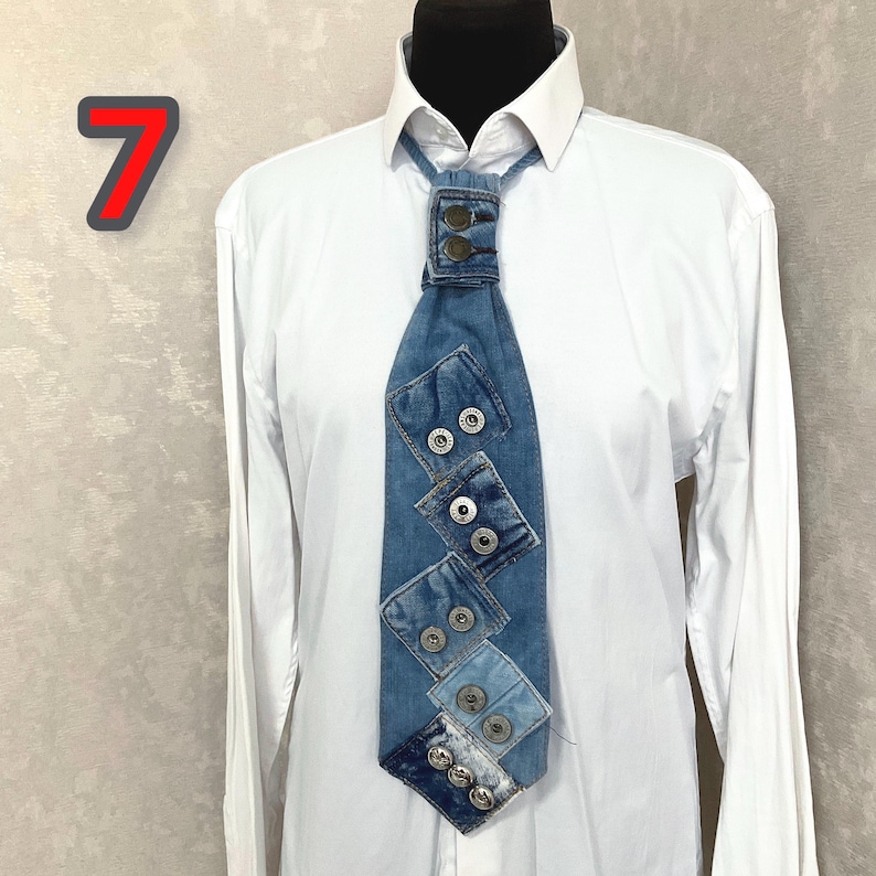 May include: A necktie crafted from blue denim fabric featuring a patchwork design with multiple silver-toned metal buttons arranged in pairs along the length. The tie is displayed on a white collared shirt with the number 7 visible in the corner.