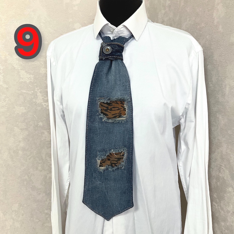 May include: A blue denim necktie featuring two distressed rectangular cutouts that reveal a brown and black animal print fabric underneath. The tie is secured at the collar with a single button and is worn over a crisp, plain white long-sleeve shirt.
