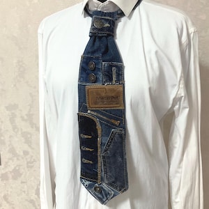 May include: A necktie constructed from repurposed blue denim fabric, featuring a brown leather patch with the text Meltin'Pot, metal buttons, a small zipper, and visible stitching details. The tie is worn over a crisp, plain white long-sleeve shirt.